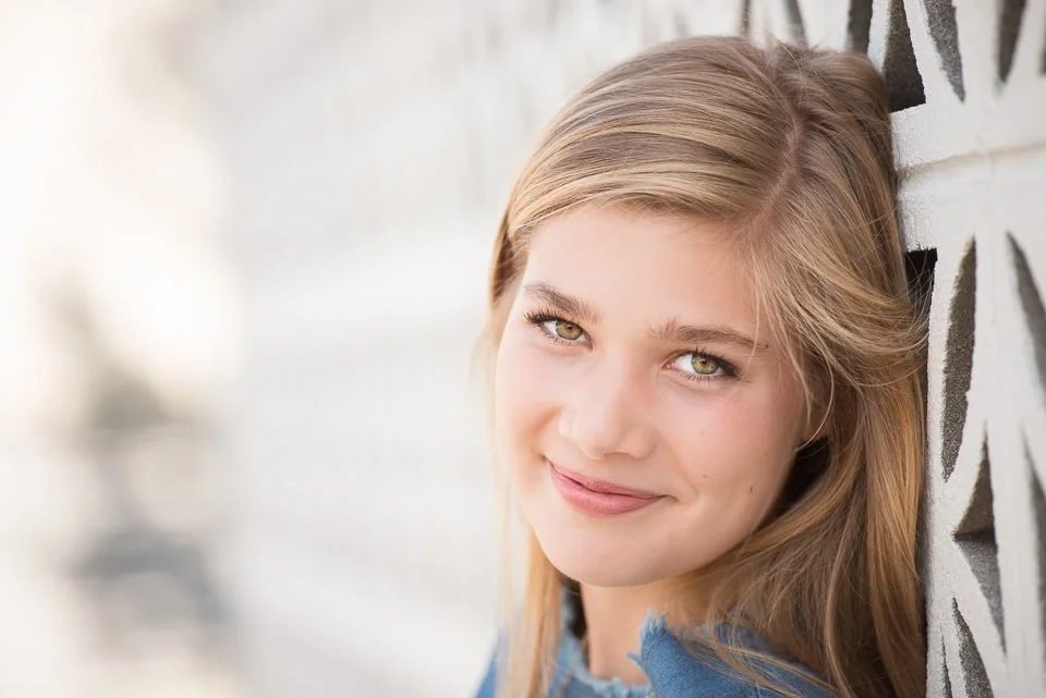 Close-up of a young woman with blonde hair and green eyes smiling and leaning against a patterned wall.