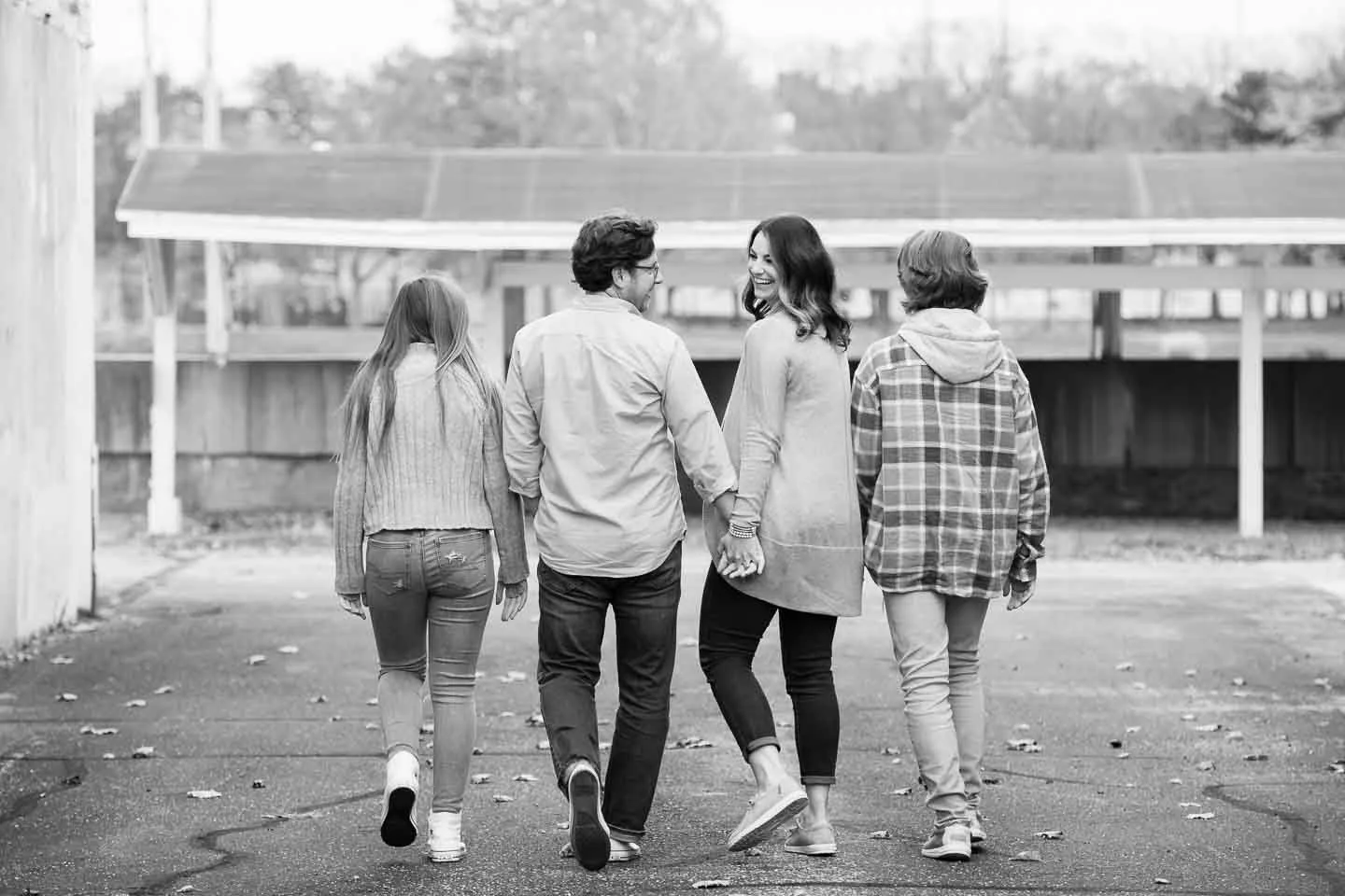 Four young people walking together outdoors, holding hands, and smiling. They are seen from the back, walking along a paved path in a park-like setting.