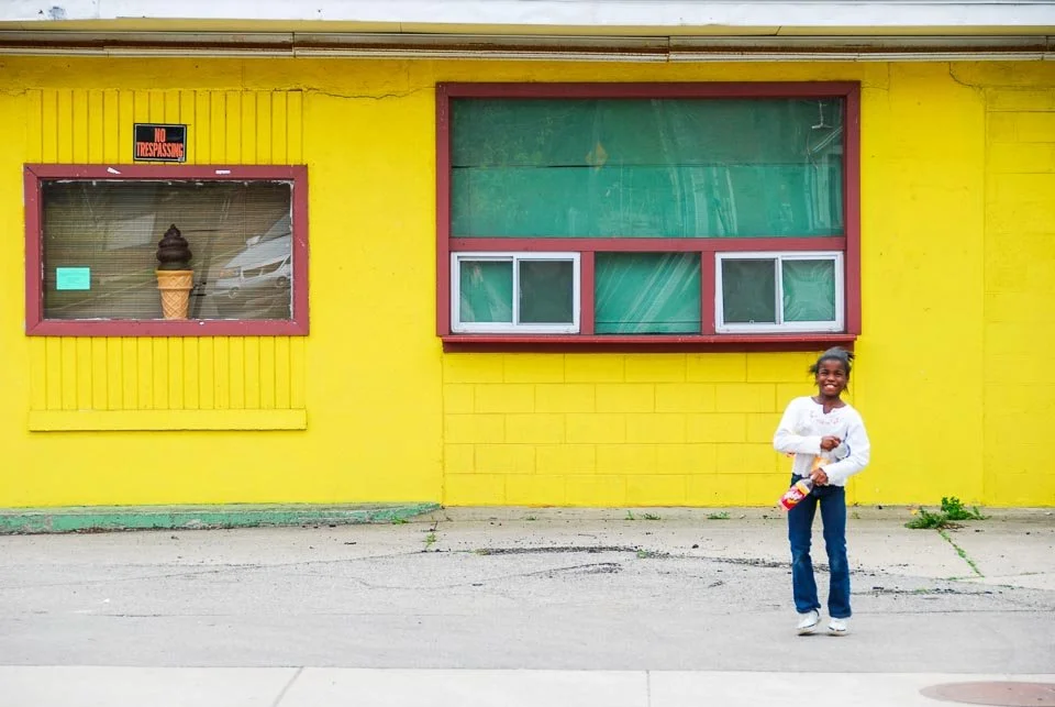 A young girl standing in front of a bright yellow building with two windows, one of which has a large ice cream cone decoration.