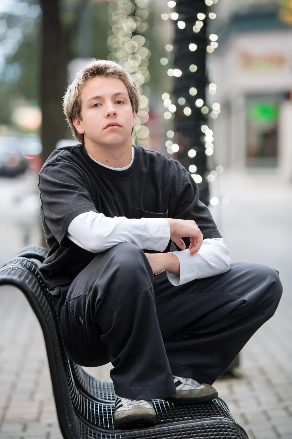 A young man with light brown hair and fair skin sitting on a black metal park bench outdoors, with blurred decorative string lights and trees in the background.