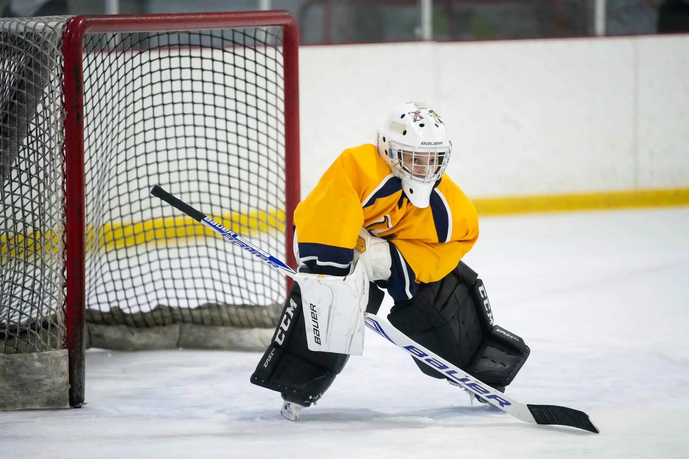 A young ice hockey goalie in a yellow jersey and white helmet, crouched in front of the net with a hockey stick, prepared to block a shot.