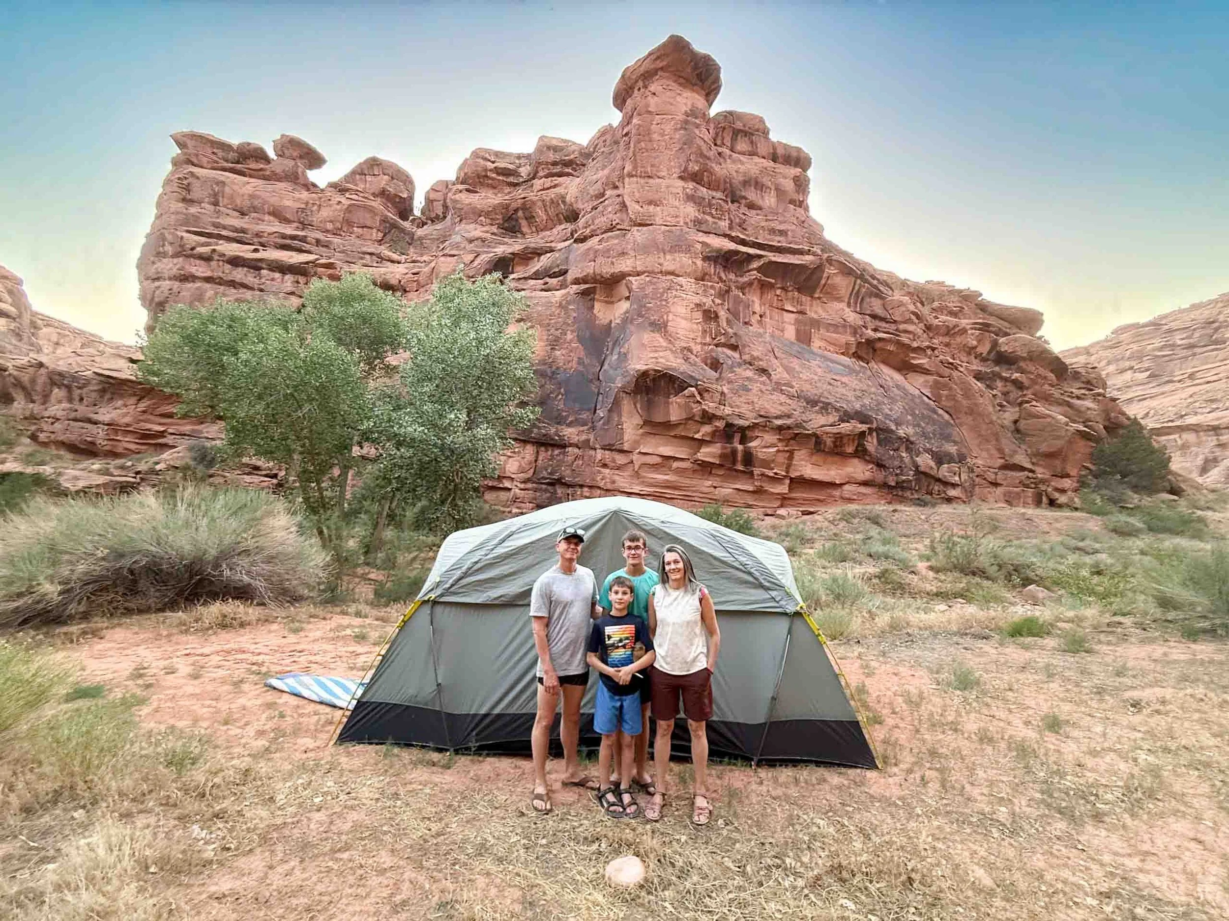 Family of four stands in front of a camping tent with red rock formations and sparse desert vegetation in the background.
