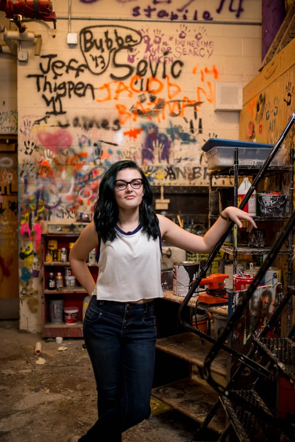 Young woman with black hair and glasses standing in an art studio with graffiti-covered walls, surrounded by painting supplies and tools.