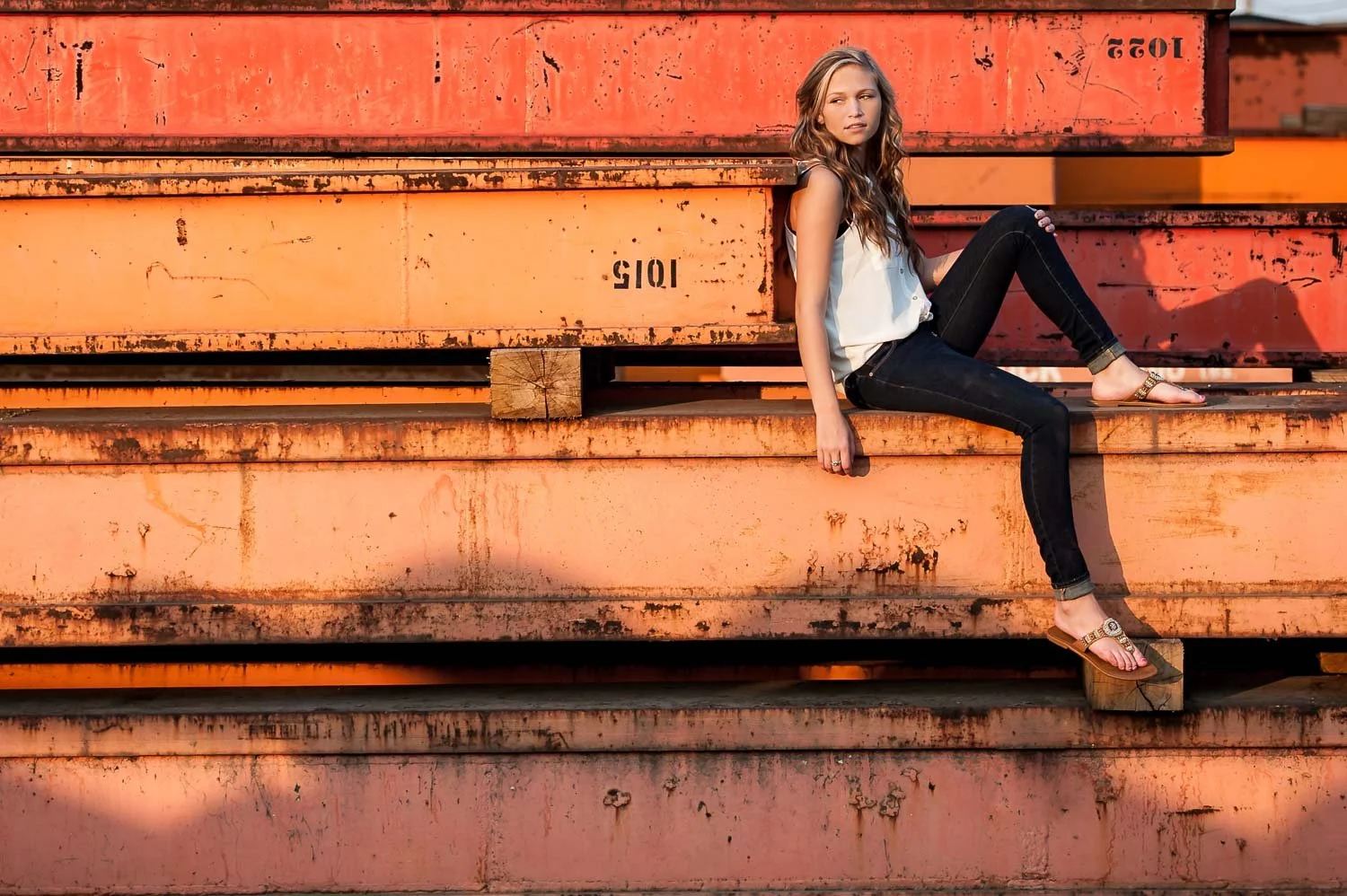 Young woman sitting on large, orange, rusted metal containers outdoors during sunset.