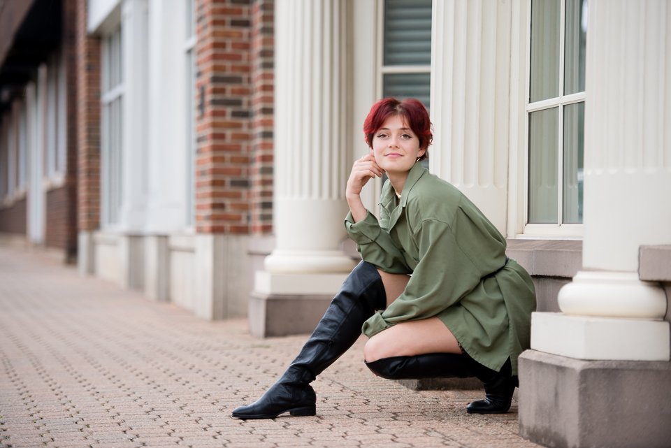 A young woman with short red hair squatting on a brick sidewalk outside a building with white columns and large windows, wearing a green jacket and black knee-high boots.
