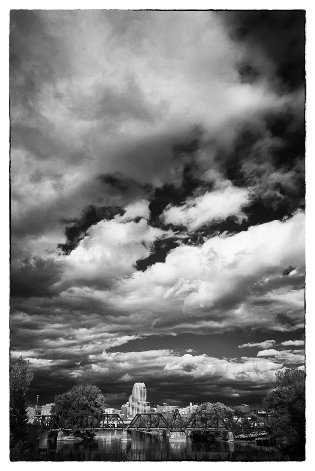 Black and white photo of a city skyline with a bridge over a body of water and dramatic cloudy sky above.