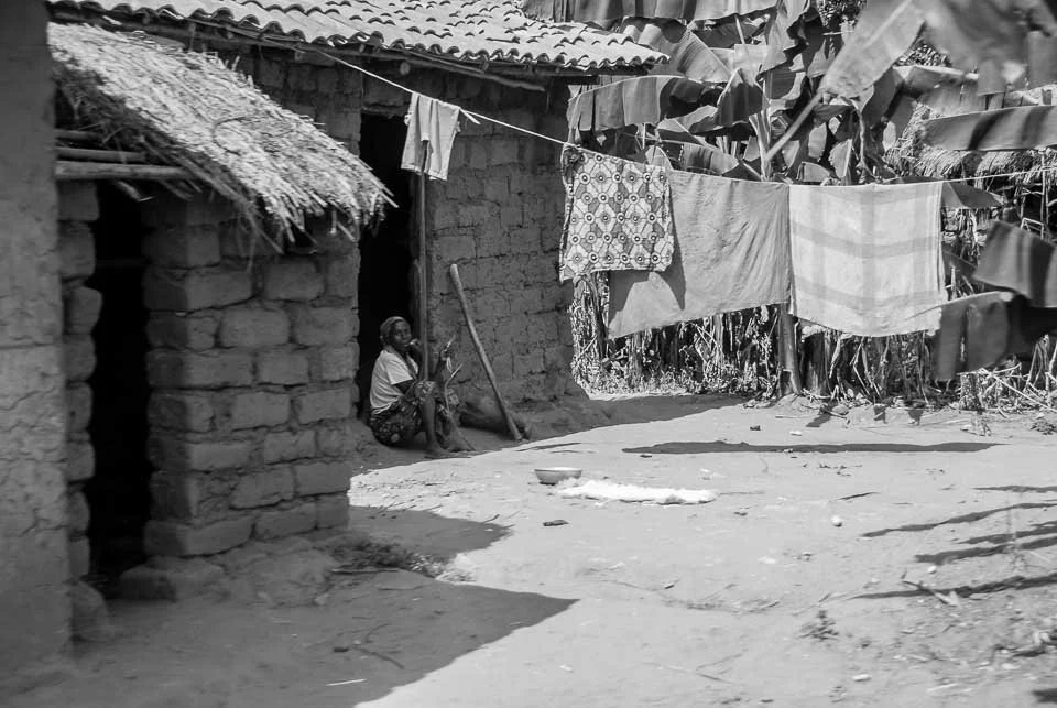 A woman sitting outside a rural hut, with clothes hanging on a line to dry in a tropical setting with banana trees in the background.