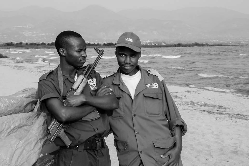 Two young men in military uniforms standing on a beach with waves and mountains in the background, one holding a rifle.