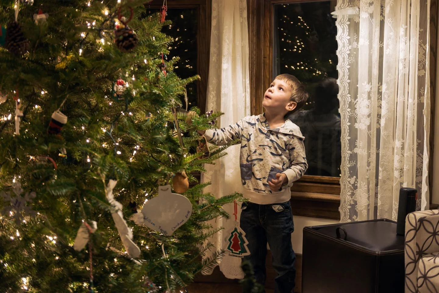 A young boy in a patterned hoodie decorating a Christmas tree with ornaments in a warmly lit room, with lace curtains and a window showing reflection of Christmas lights.