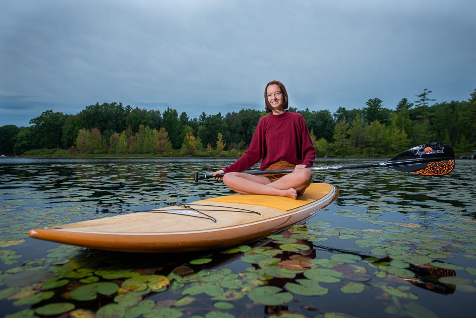 A woman sitting cross-legged on a paddleboard holding a paddle on a lake surrounded by lily pads and trees, with a cloudy sky overhead.