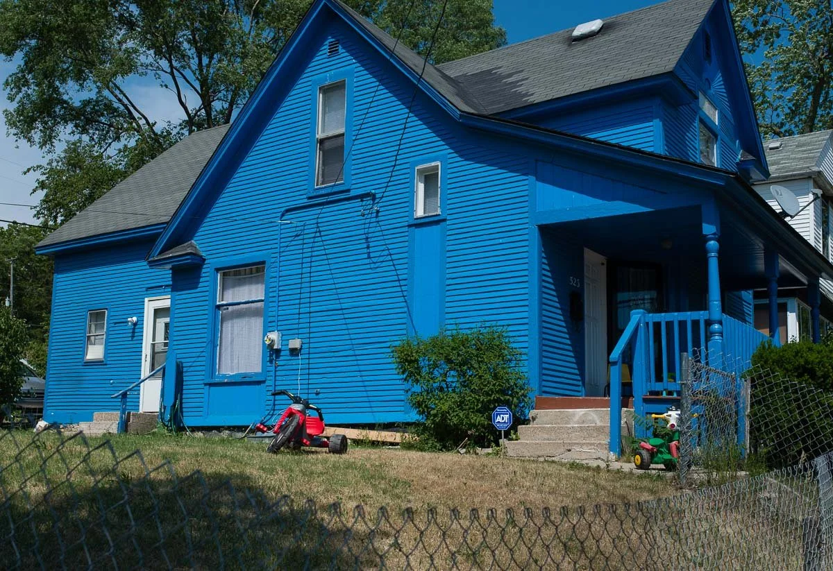 A bright blue two-story house with a front porch and steps leading up to the door. There is a small bush near the steps, a chain-link fence in the front yard, and children's toys on the lawn, including a red push lawnmower and a green and red tricycl