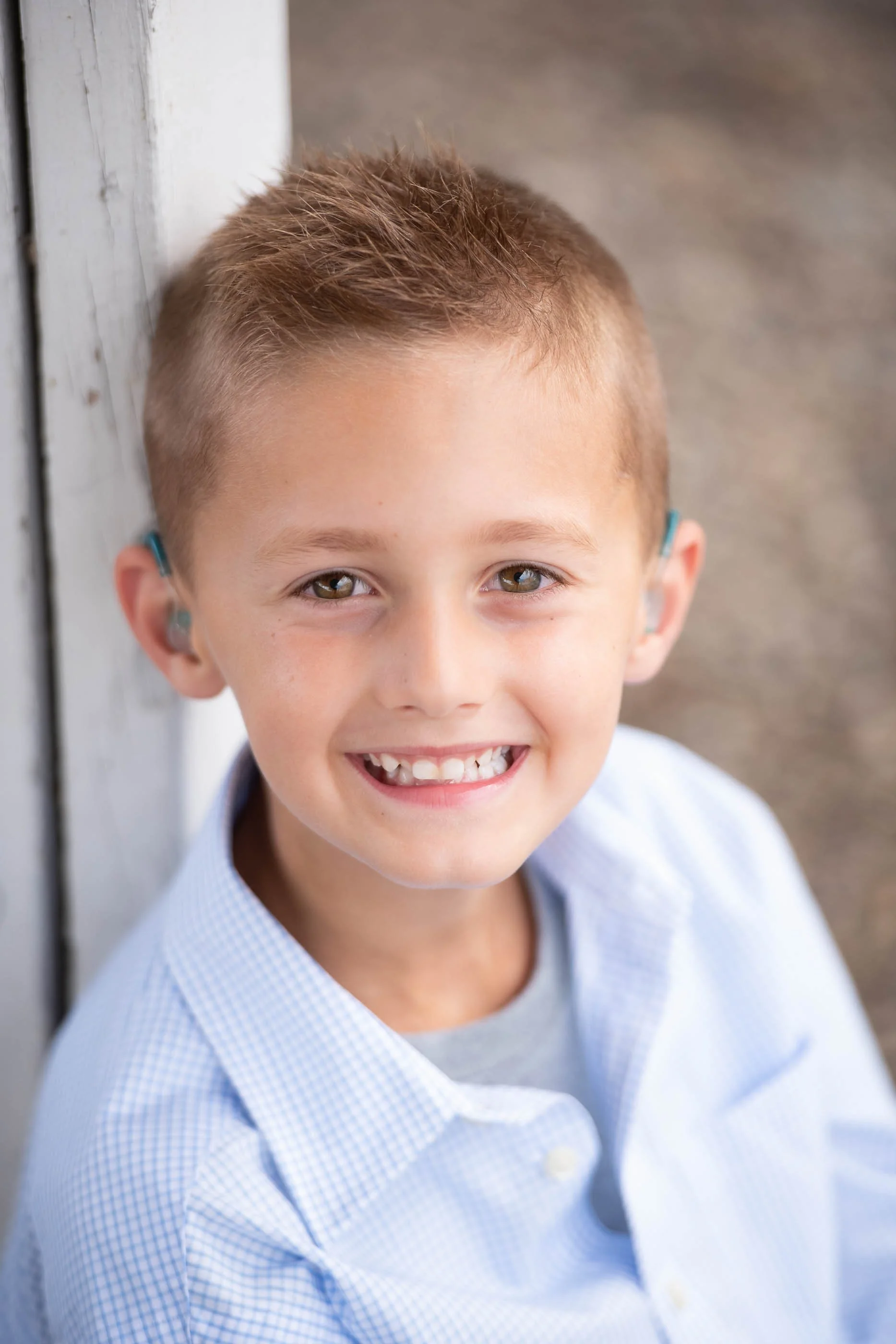 Young boy with light brown hair, wearing a light blue checkered shirt, smiling, and sitting outdoors near a wooden structure.