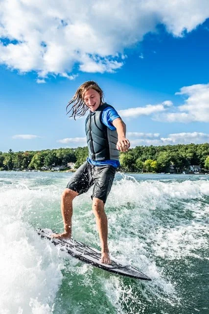 A young boy surfing on a river, wearing a life jacket and shorts, under a partly cloudy sky.