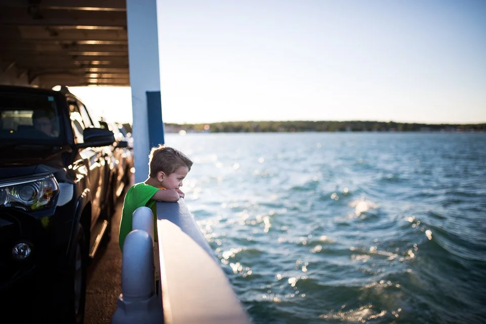 A young boy leaning on the railing of a ferry boat, looking out at a large body of water during daylight.