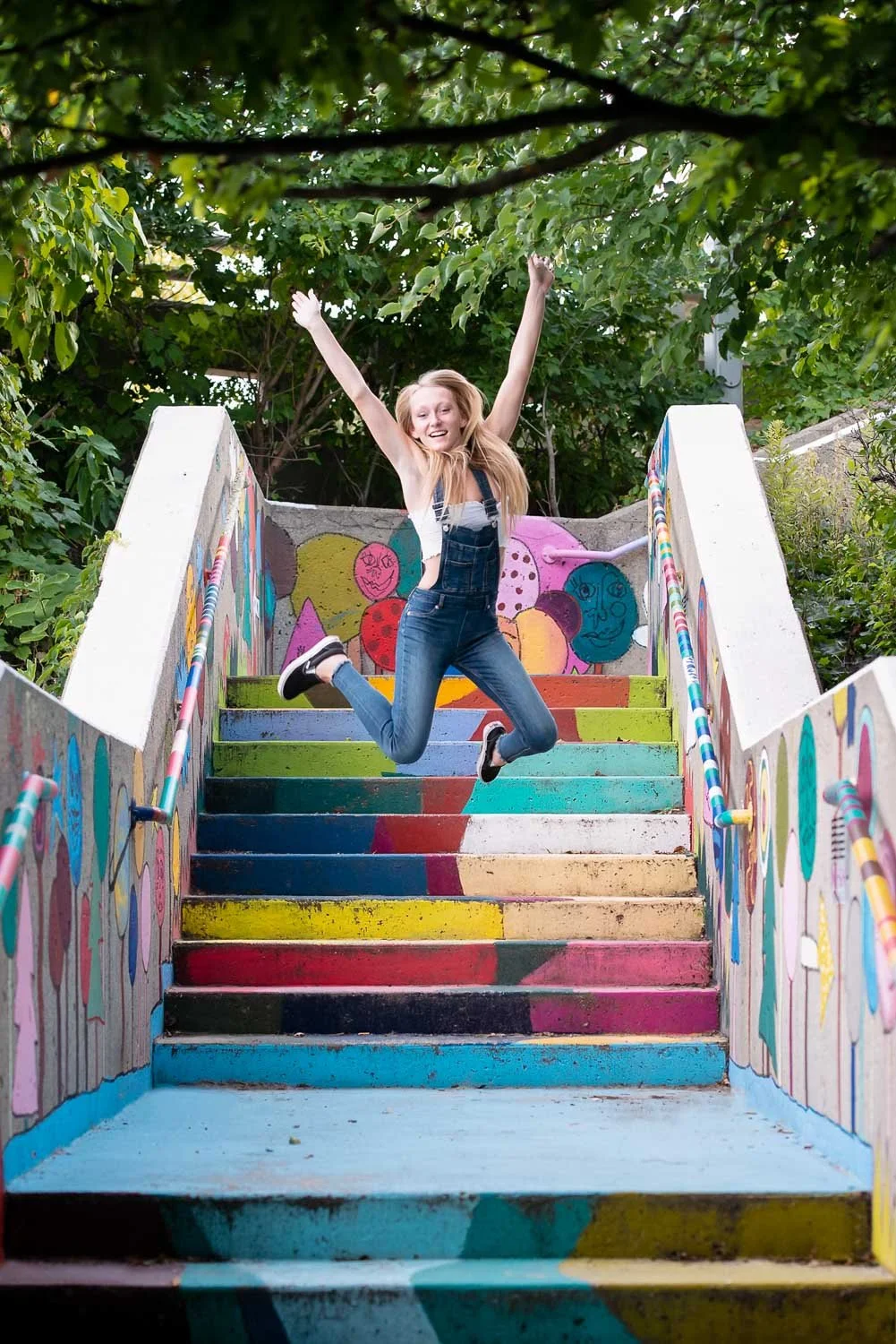 A young woman with long blonde hair jumping in the air on colorful, graffiti-decorated stairs outdoors surrounded by green trees.
