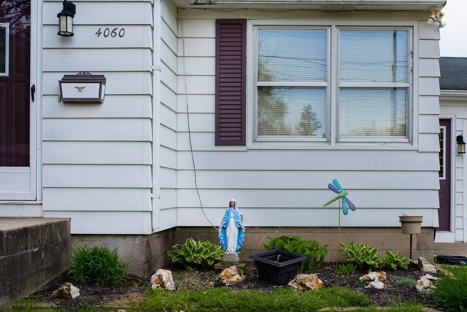 Front yard of a house with a religious figurine of the Virgin Mary, a decorative dragonfly, plant pots, and landscaped plants.