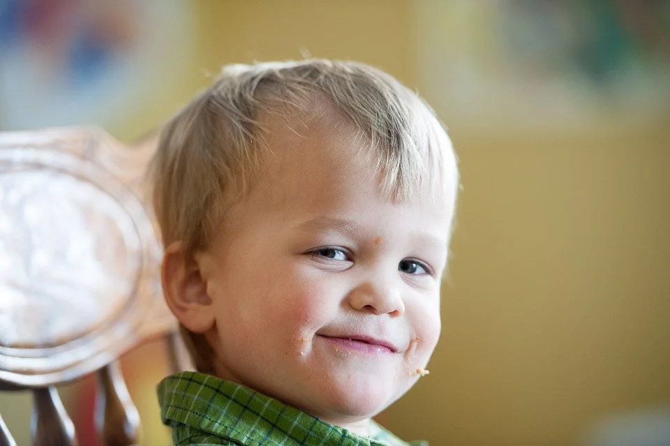 A young boy with short blond hair and fair skin has a slight smile and is looking at the camera, with food around his mouth.