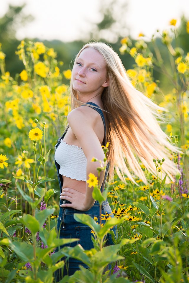 A young woman with long blonde hair standing in a field of yellow flowers during daylight, looking over her shoulder with a slight smile.