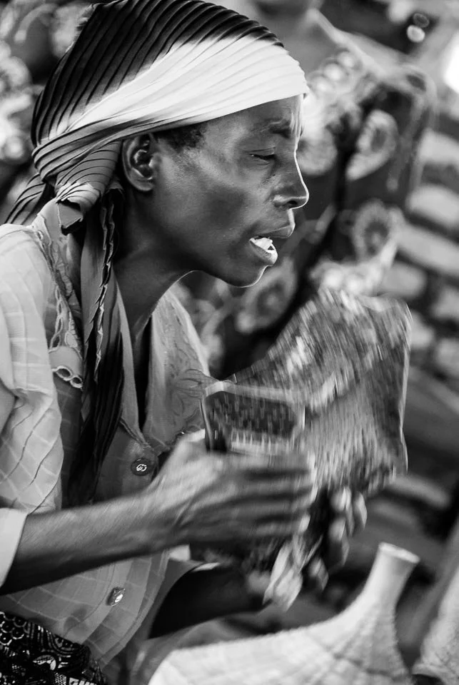 Black and white photo of a woman wearing a striped headscarf, holding and looking at a newspaper.