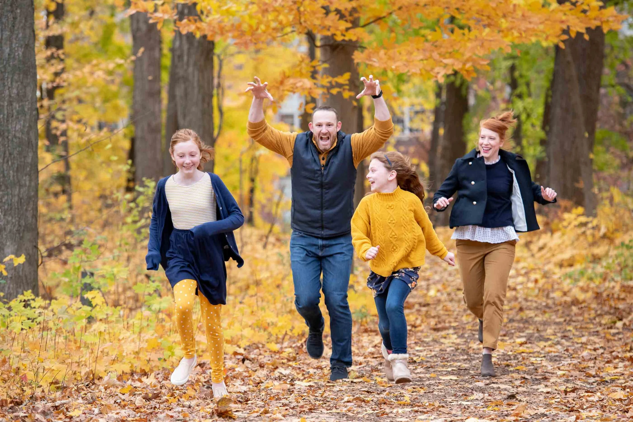 A group of five people, one adult male and four young girls, running and laughing in an autumn forest with yellow and orange leaves.