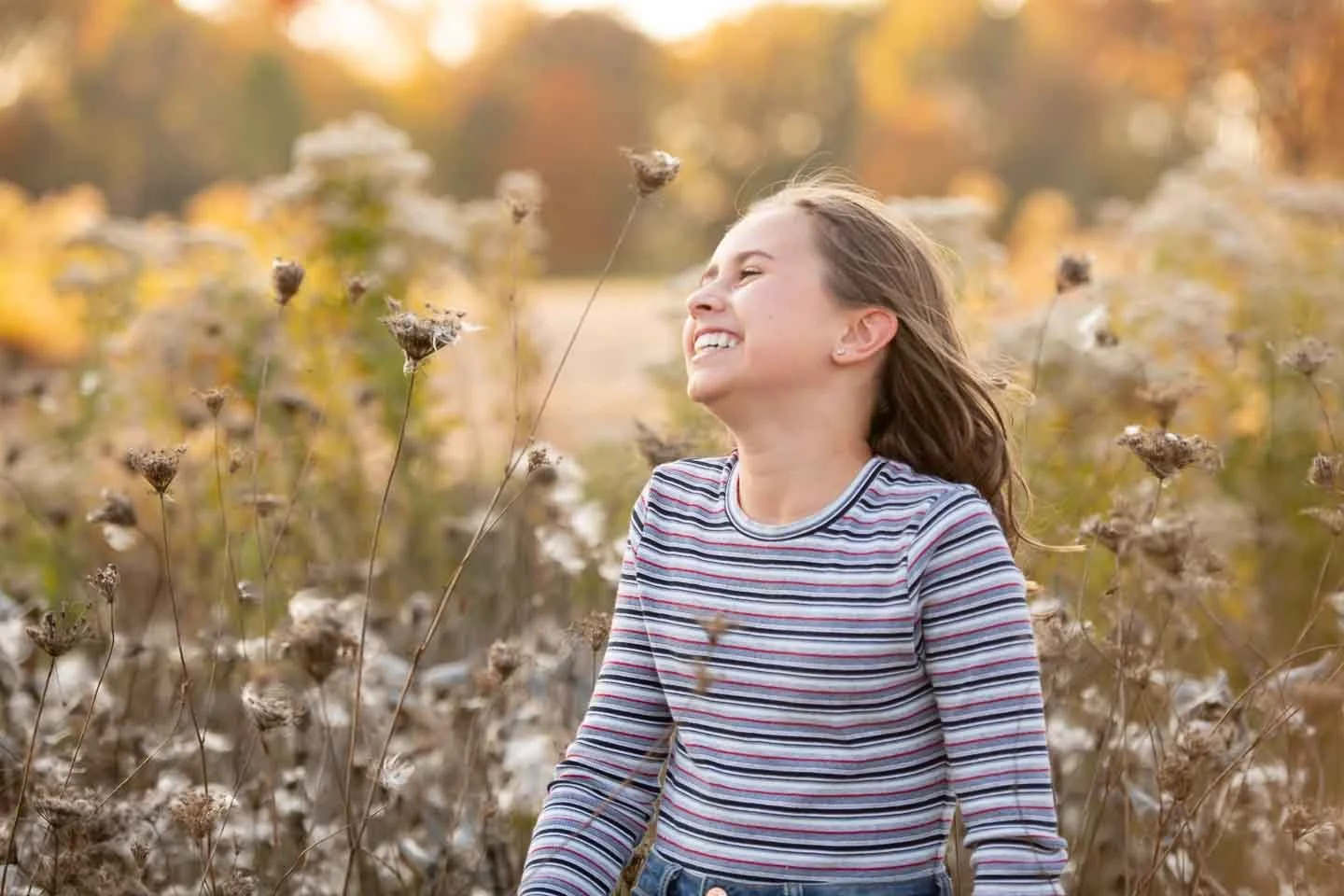 A girl with long brown hair smiling and enjoying herself in a field of dry plants with autumn-colored trees in the background at sunset.
