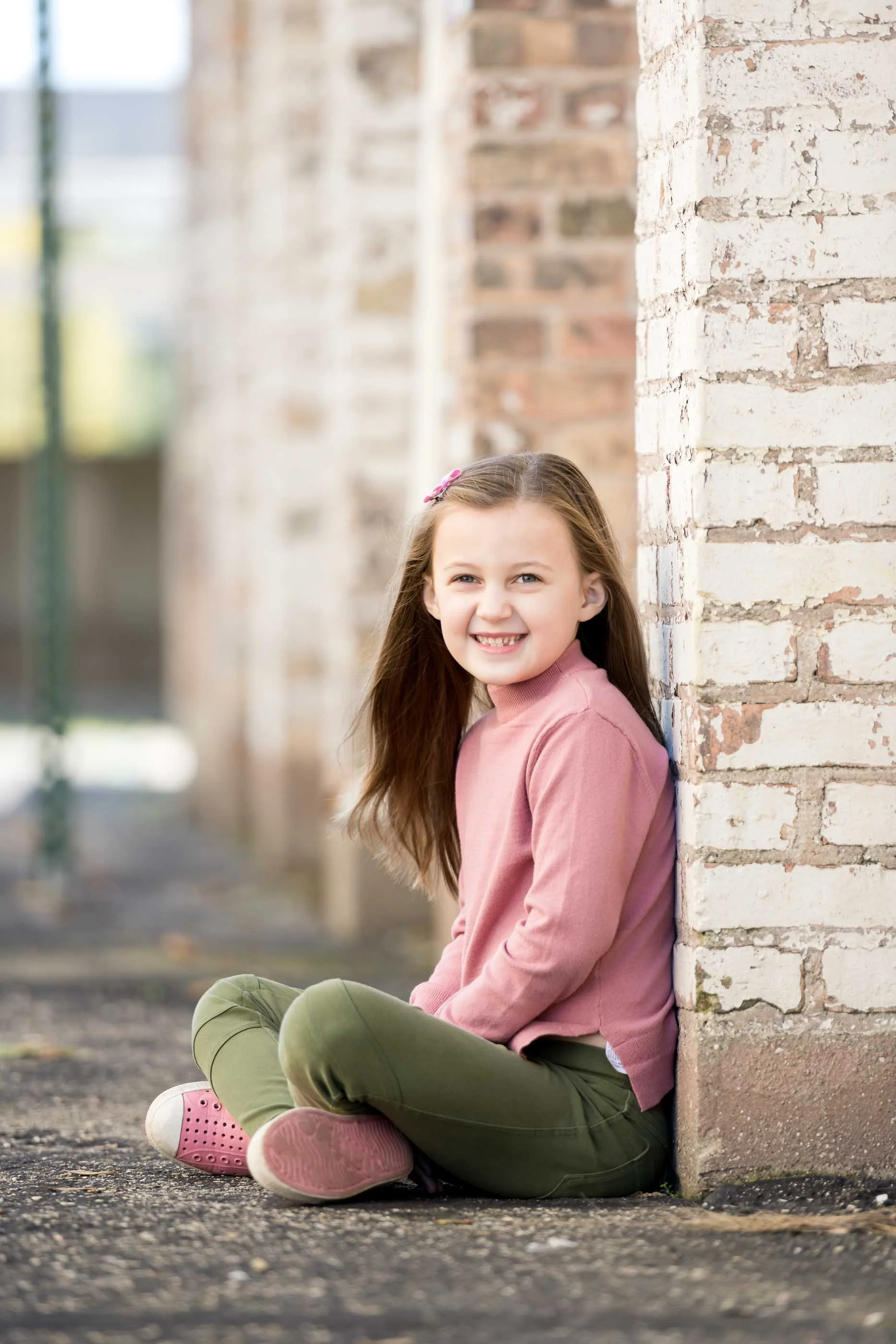A young girl with long brown hair, wearing a pink sweater, olive green pants, and pink shoes, sitting on the ground and smiling next to a brick wall.