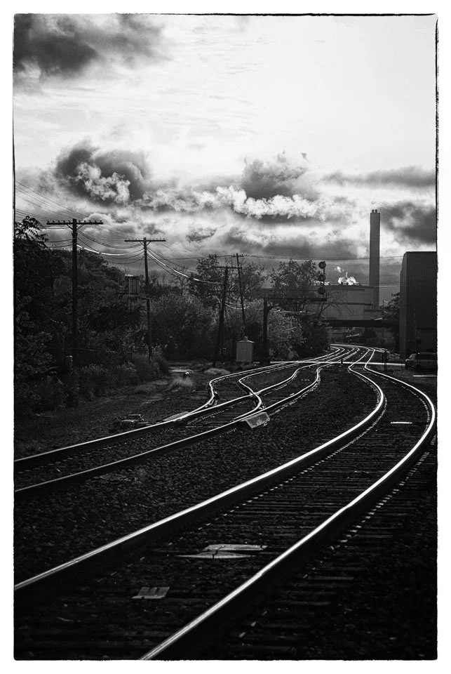 Black and white photo of railroad tracks curving into the distance with cloudy sky overhead.