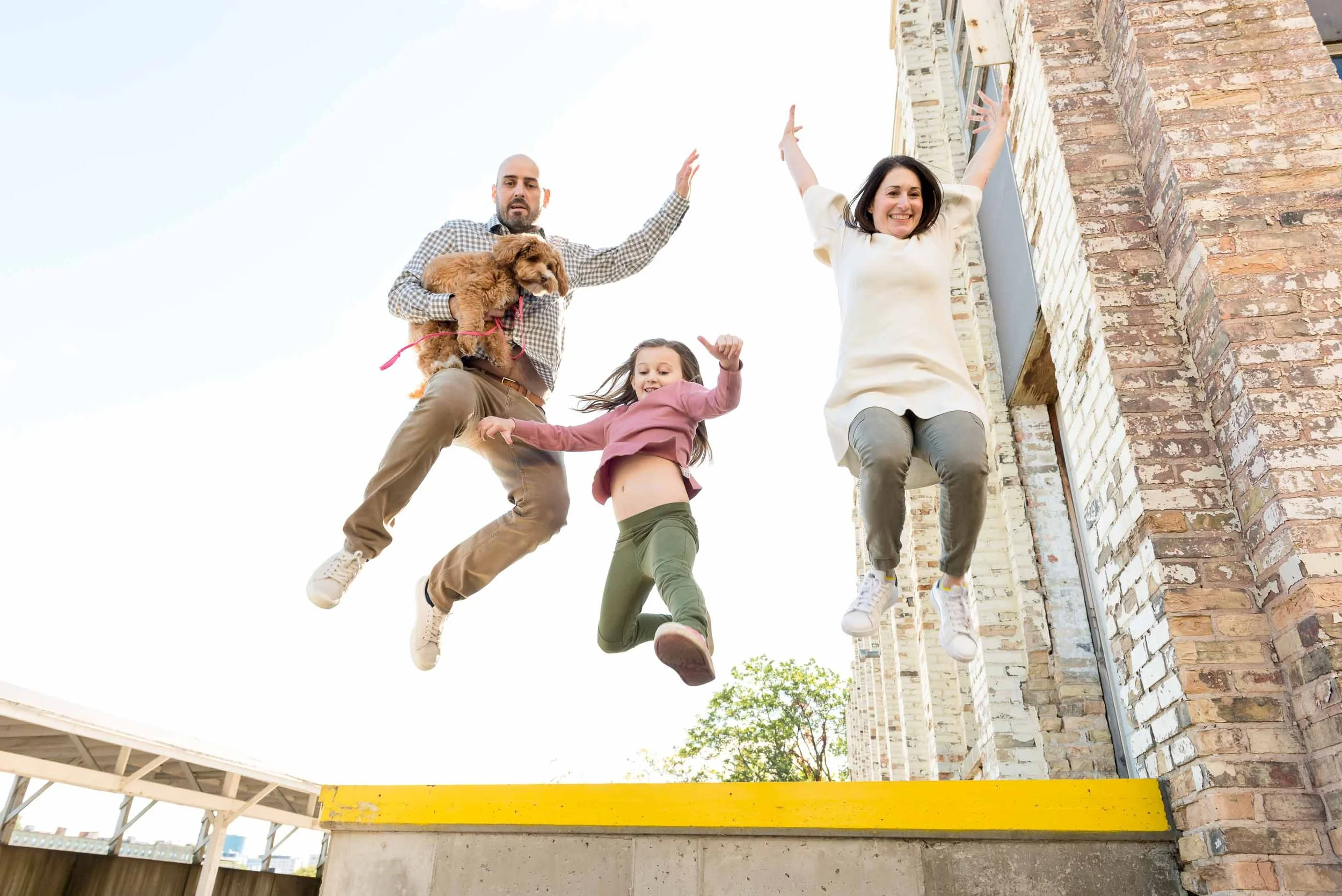 A family of four and a dog jumping in the air outdoors in front of a brick building, with a clear blue sky above.