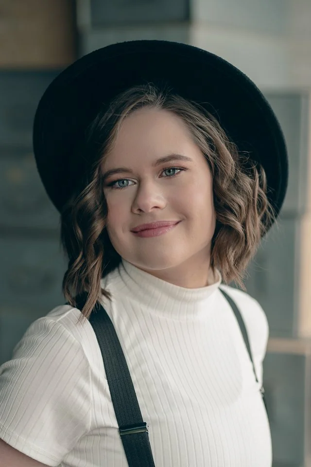 A young woman with wavy brown hair wearing a black hat, white ribbed turtleneck, and black suspenders.