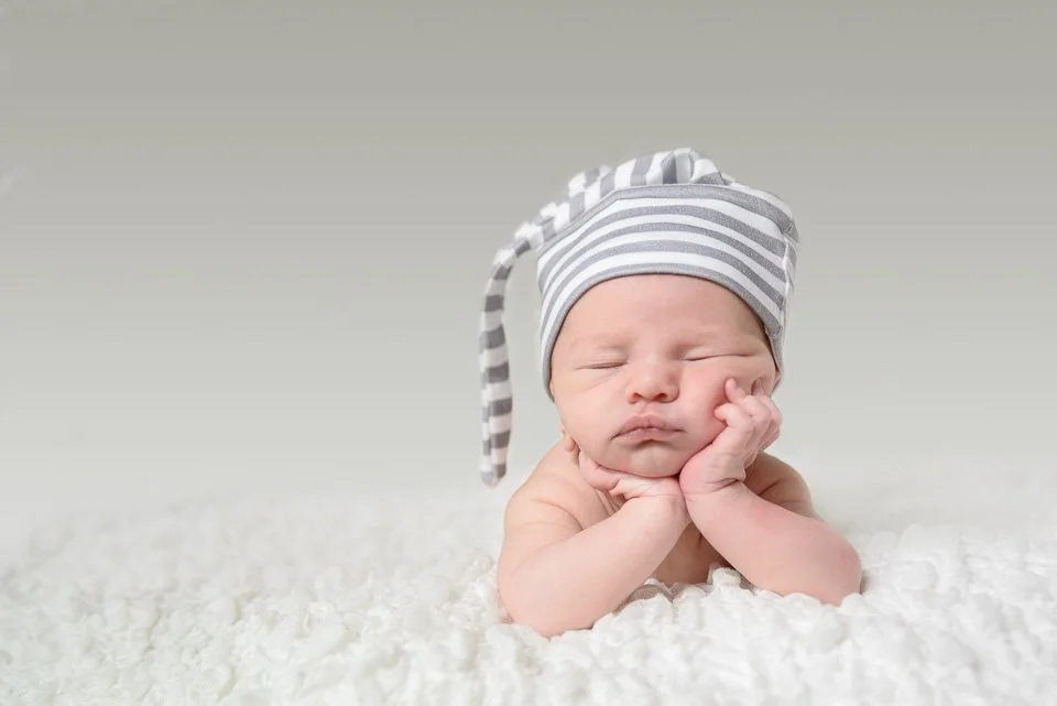 A sleeping baby lying on a soft, white blanket, with a gray and white striped hat on its head.