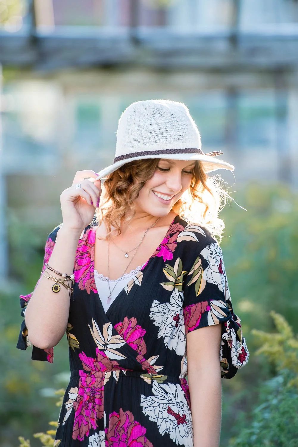 A woman with curly hair wearing a floral dress and white hat, smiling outdoors.
