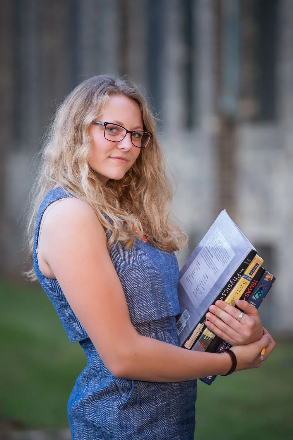 A young woman with curly blonde hair and glasses holding a stack of books outdoors, looking at the camera.