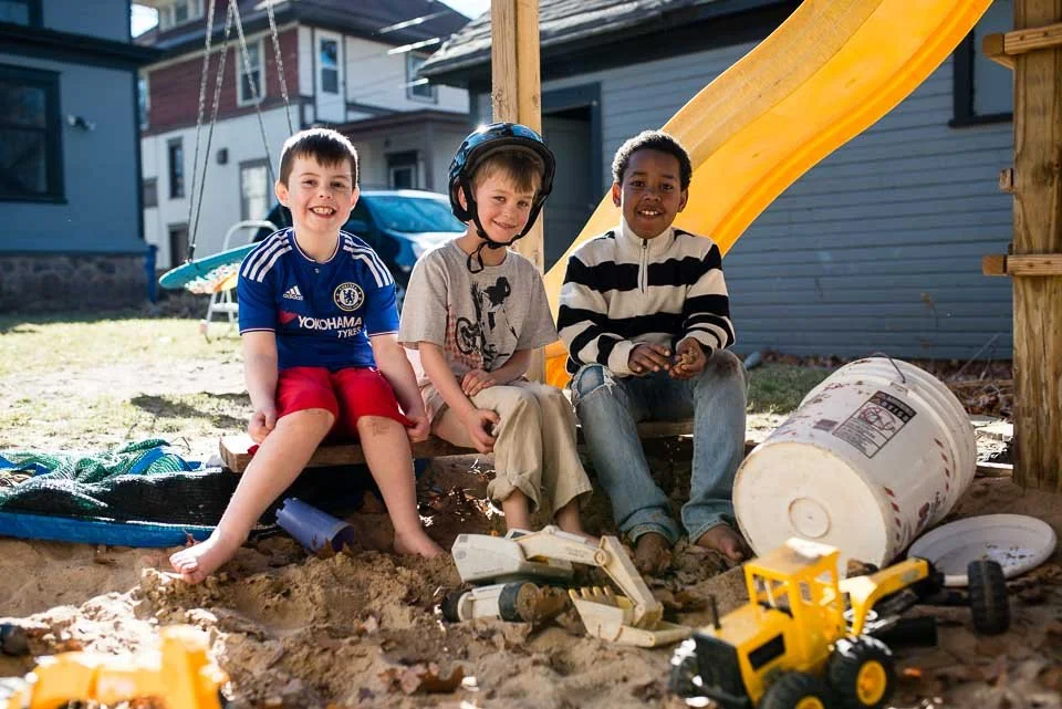 Three children playing on a sandbox with a slide and various toys, smiling at the camera.