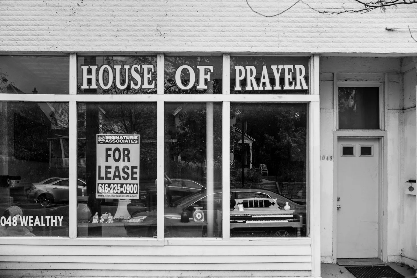Front of a building with large window display reading 'House of Prayer' and a 'For Lease' sign in the window, reflecting parked cars outside and trees.