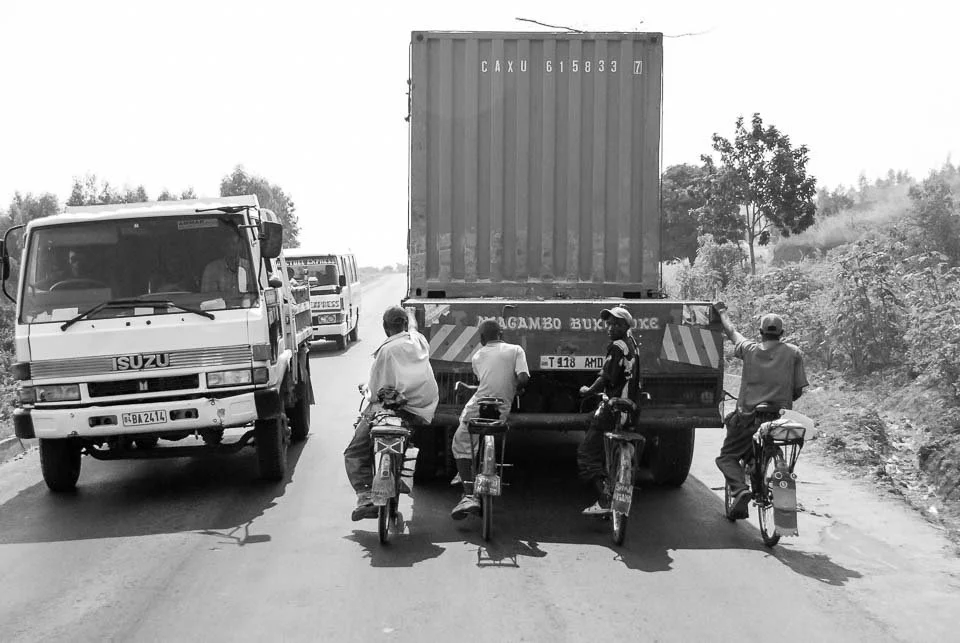 People riding bicycles on a road in front of a large truck, with a pickup truck and another vehicle in the background.