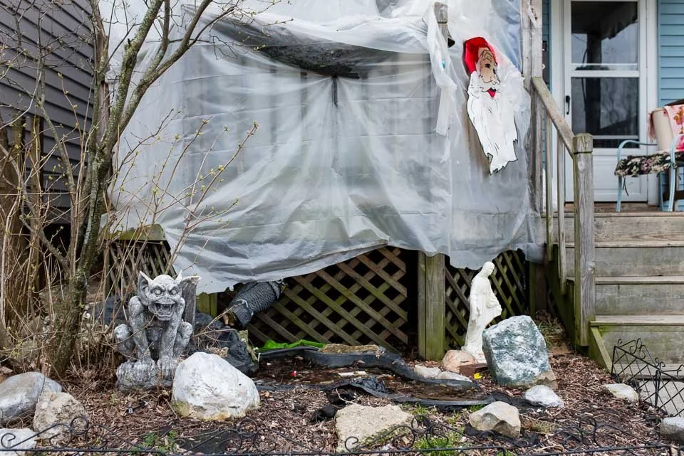 Decorations for Halloween outside a house, including a Santa face hanging near the porch, a stone gargoyle, a ghost figure, and rocks and plants in the yard.