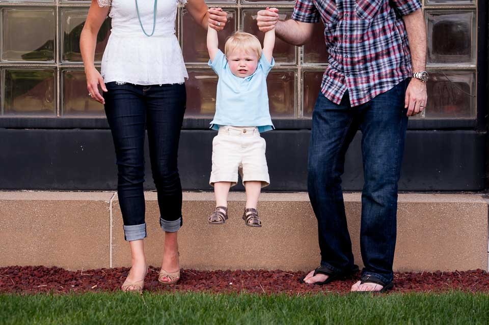 A young boy is holding hands with two adults, one on each side, while being lifted off the ground. The boy appears to be sad or upset. The adults are only partially visible, and the image is set outdoors in front of a building with glass block window
