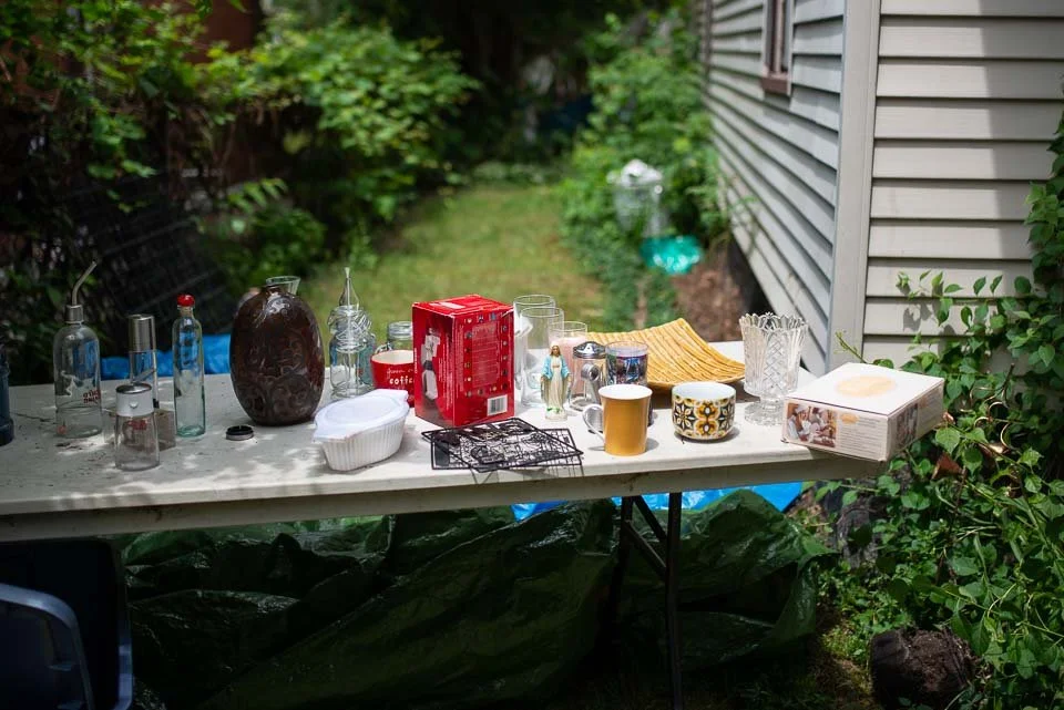 Outdoor table with various glassware, a box of cookies, and cups, set in a garden near a house with siding.