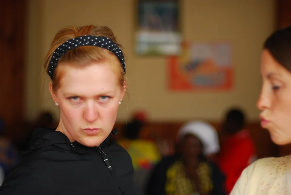 A young woman with red hair, wearing a black hoodie and a polka dot headband, looking directly at the camera with a serious expression in an indoor setting with other people in the background.
