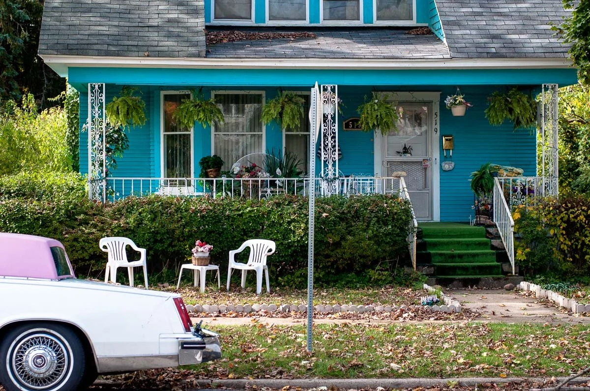 A turquoise house with white porch railing and hanging plants, green steps leading to the front door, and a vintage car with a pink roof parked in front.