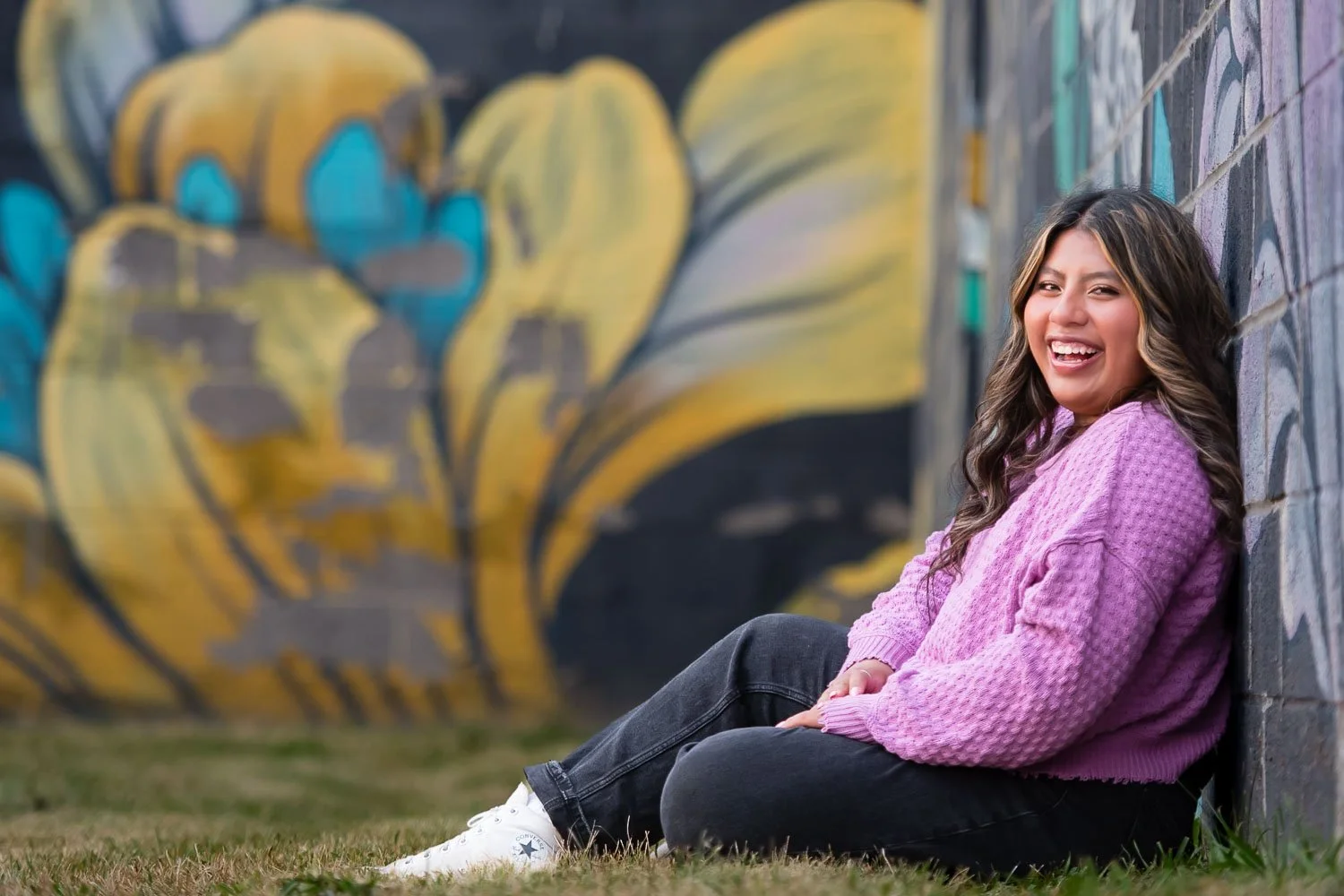 A young woman with long, wavy hair sitting on grass against a graffiti-covered brick wall, smiling and looking at the camera. She is wearing a pink sweater and white sneakers, with a colorful mural behind her.