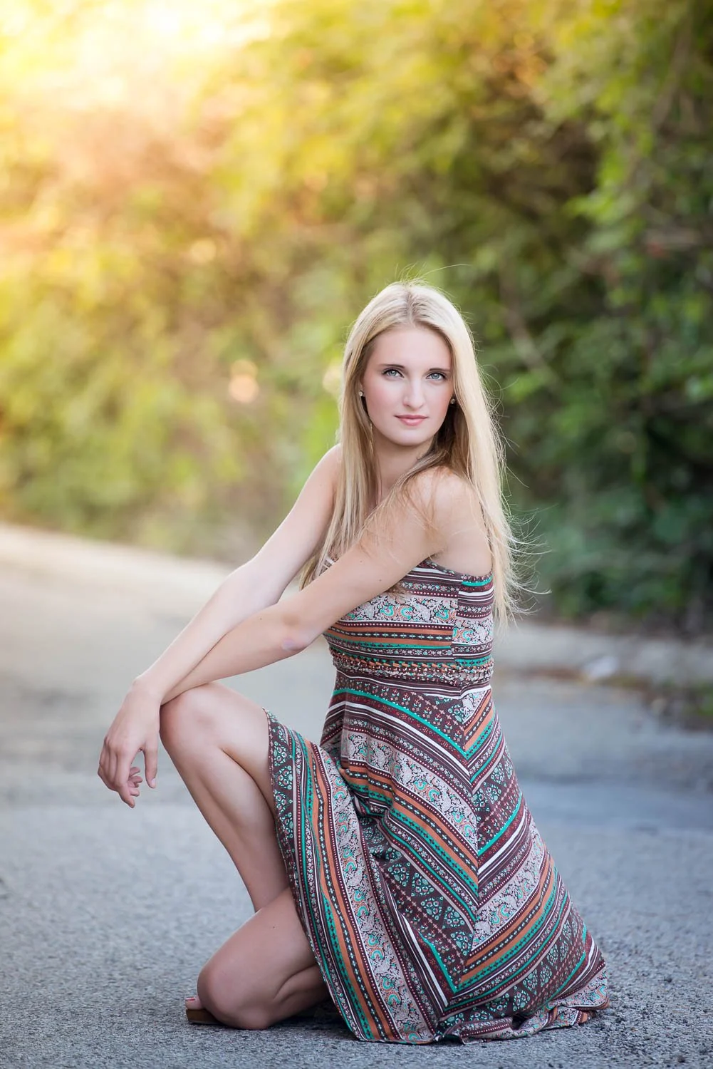 A young woman with long blonde hair in a patterned dress, kneeling on a paved outdoor path with greenery and sunlight in the background.