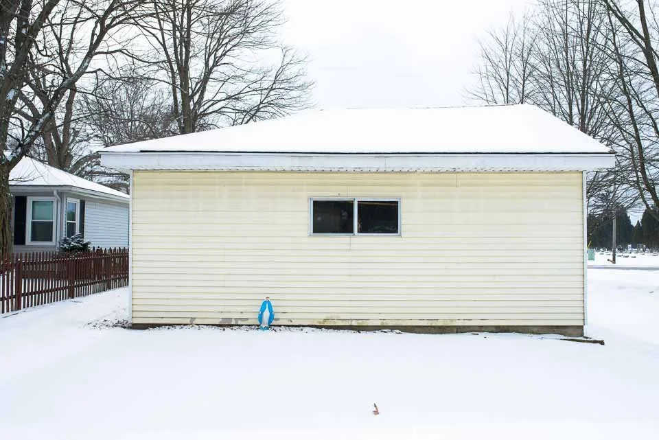 Back of a house with beige vinyl siding, snow-covered ground, and a snow-covered roof. A window near the top center and a blue toy figure near the bottom on the snow.