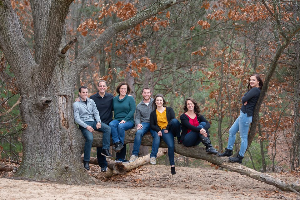 Group of seven people, three women and four men, sitting and standing on a large tree branch in a wooded area with autumn leaves.