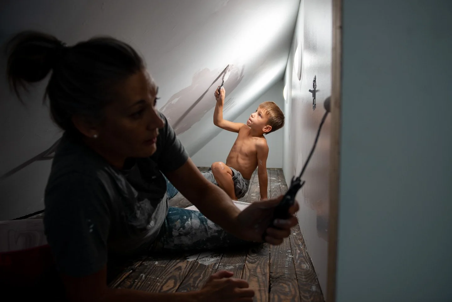 A woman painting a wall while a young shirtless boy sits on the floor, looking at her.