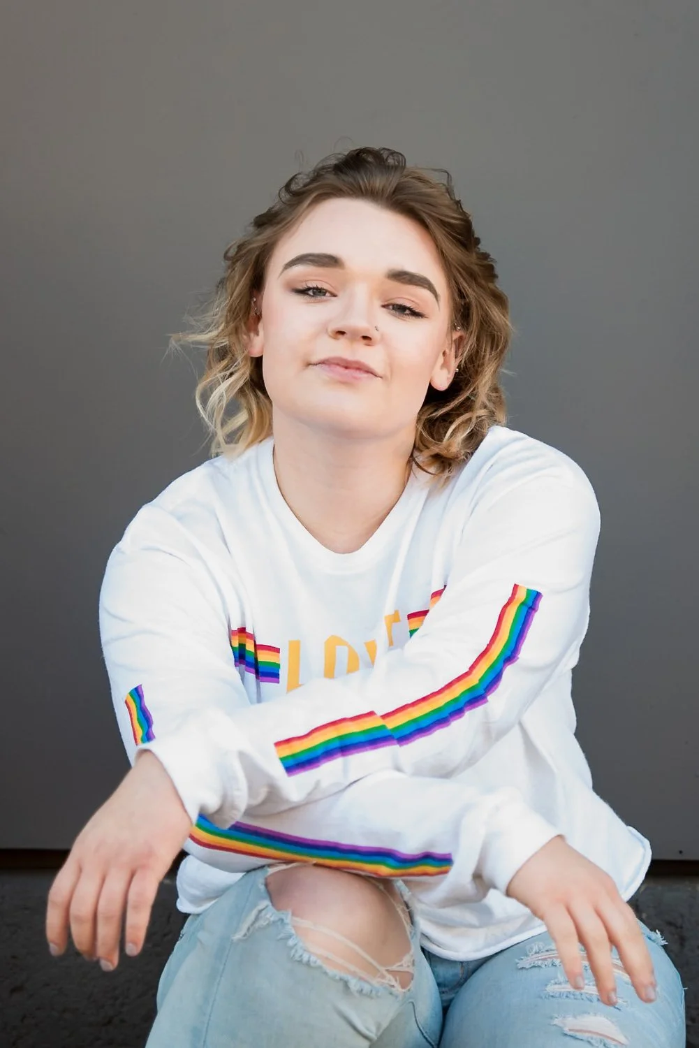 A young woman with shoulder-length blonde hair, fair skin, and makeup, sitting against a plain gray background. She is wearing a white long-sleeve shirt with rainbow stripes on the sleeves and a rainbow flag on the front, along with ripped light blue
