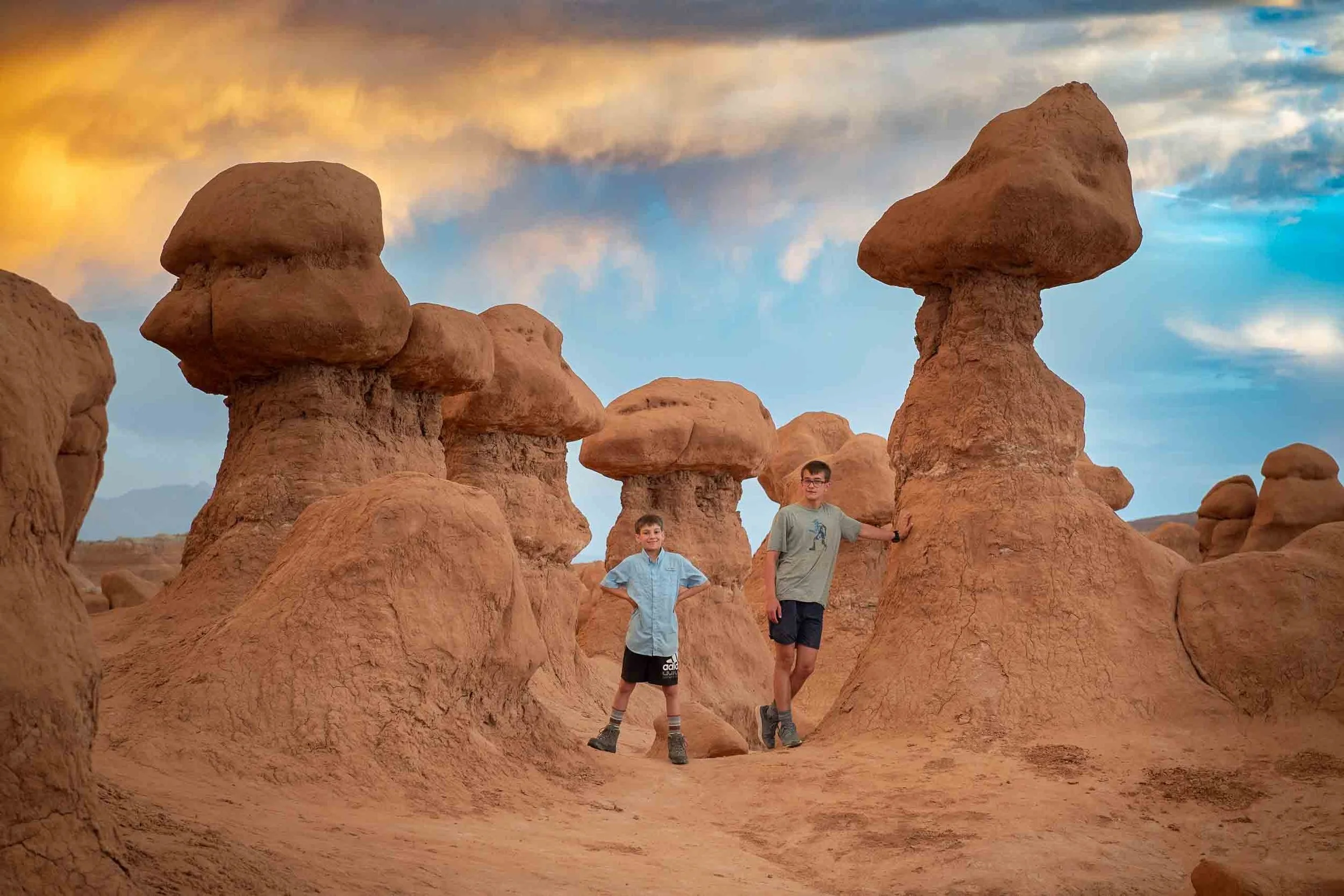 Two boys standing among unique rock formations resembling mushrooms in a desert landscape during sunset.