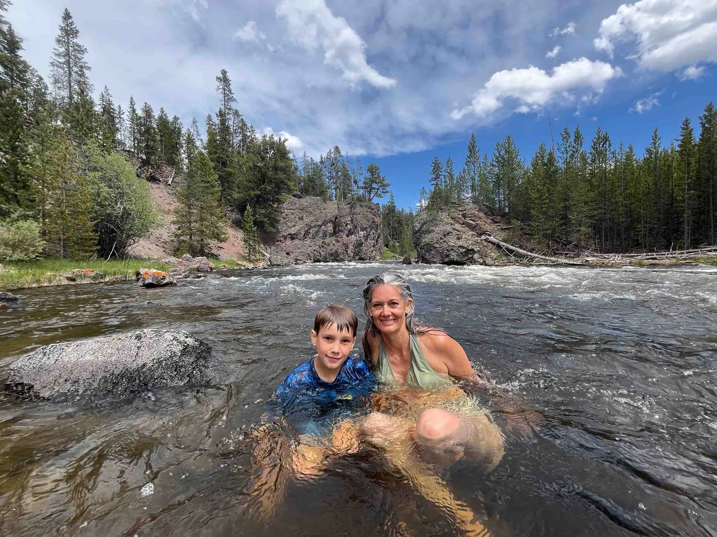 A woman and a young boy swimming in a river with a forest and rocky cliffs in the background under a partly cloudy sky.