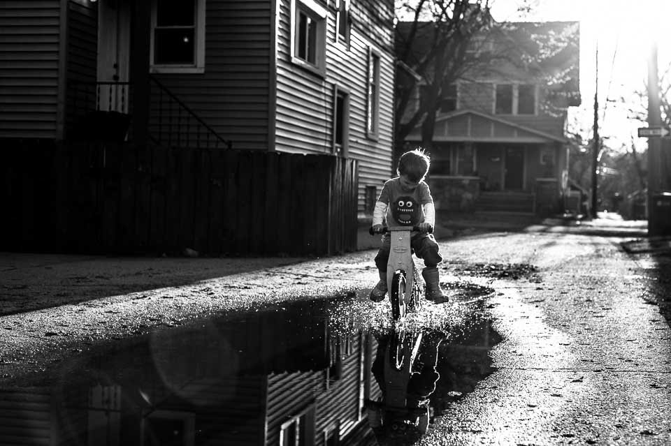 A young child riding a bike through a puddle on a residential street, with houses and trees in the background, in black and white.