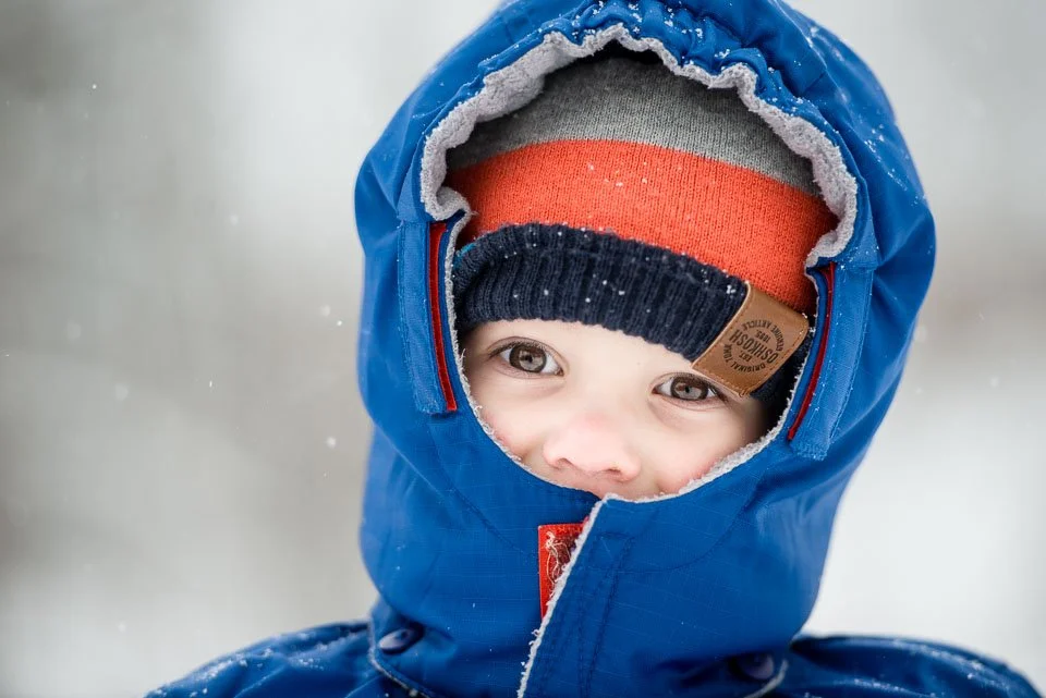 Close-up of a young child in winter clothes, smiling with smiling eyes, wearing a blue hooded winter jacket, a multicolored beanie, and a black knit hat underneath, surrounded by falling snow.